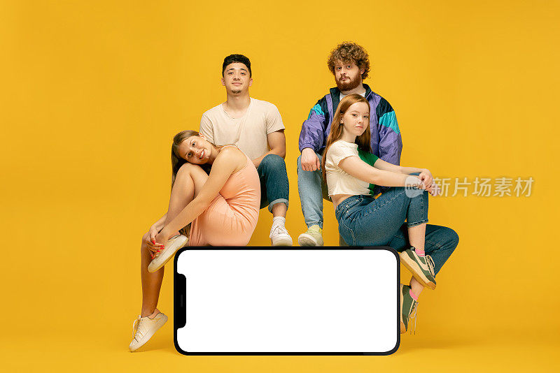 一群年轻人站着或坐着，坐在巨大的3d智能手机模型上，黄色背景上有空白的白色屏幕，网站，应用程序，techno, Mockup